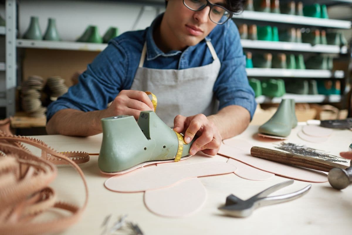 A person in an apron measures leather with a tape at a workbench, surrounded by tools and shoe forms in a workshop.