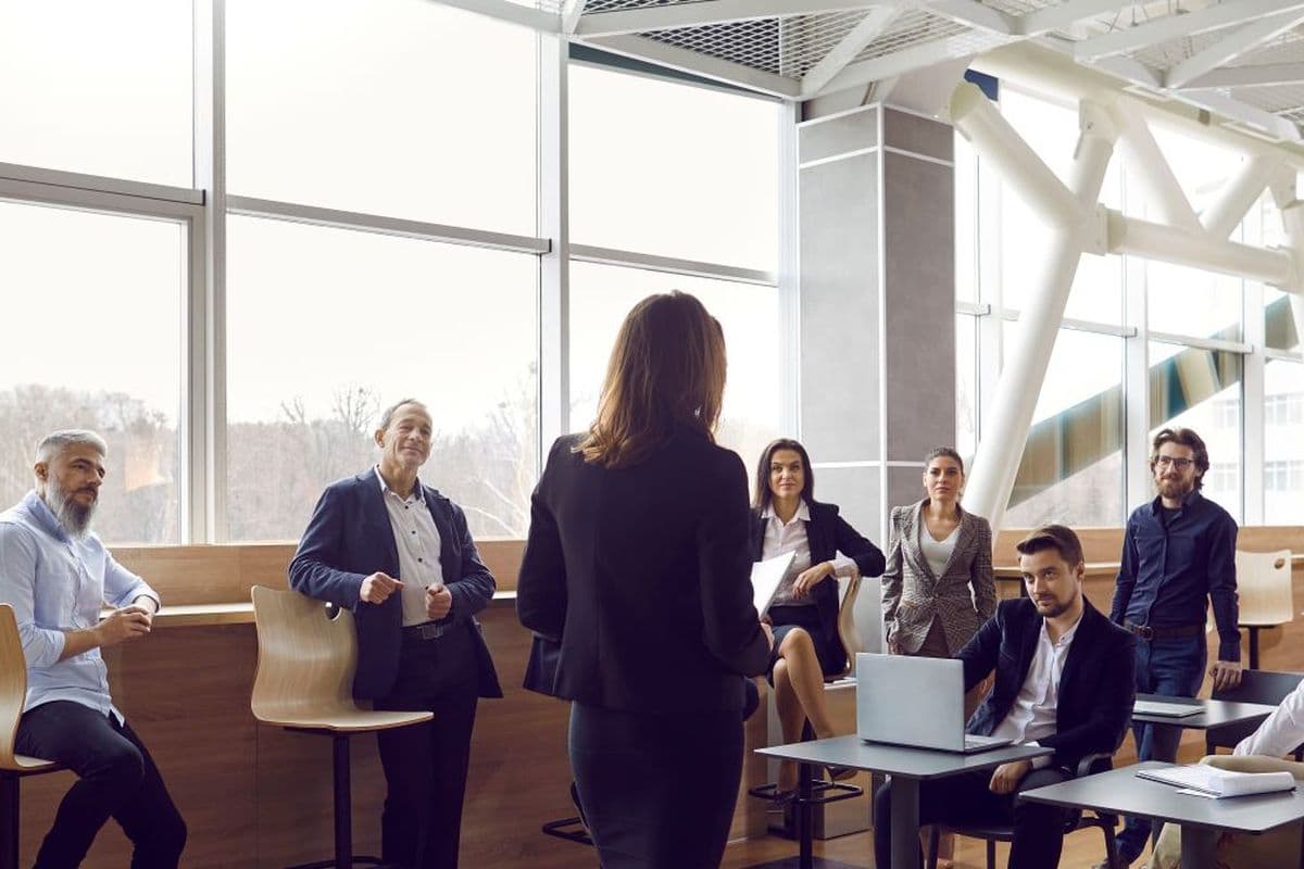 A woman stands addressing a group of seated colleagues in a modern office with large windows and natural light.