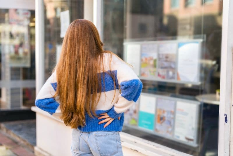 During daylight hours, a young woman examines real estate listings displayed in a shop window