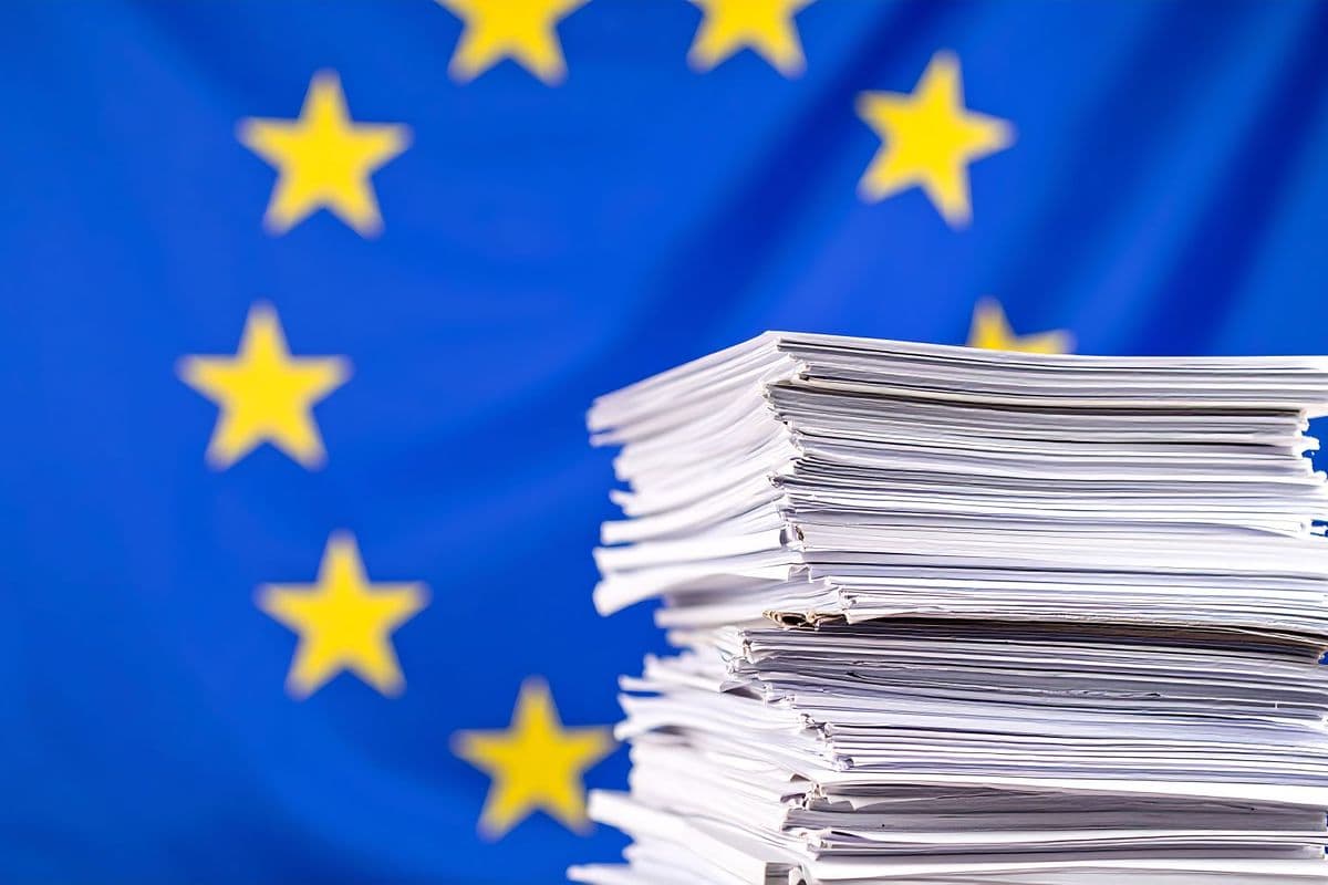 Stack of papers in front of a blurred European Union flag, symbolizing bureaucracy or documentation.