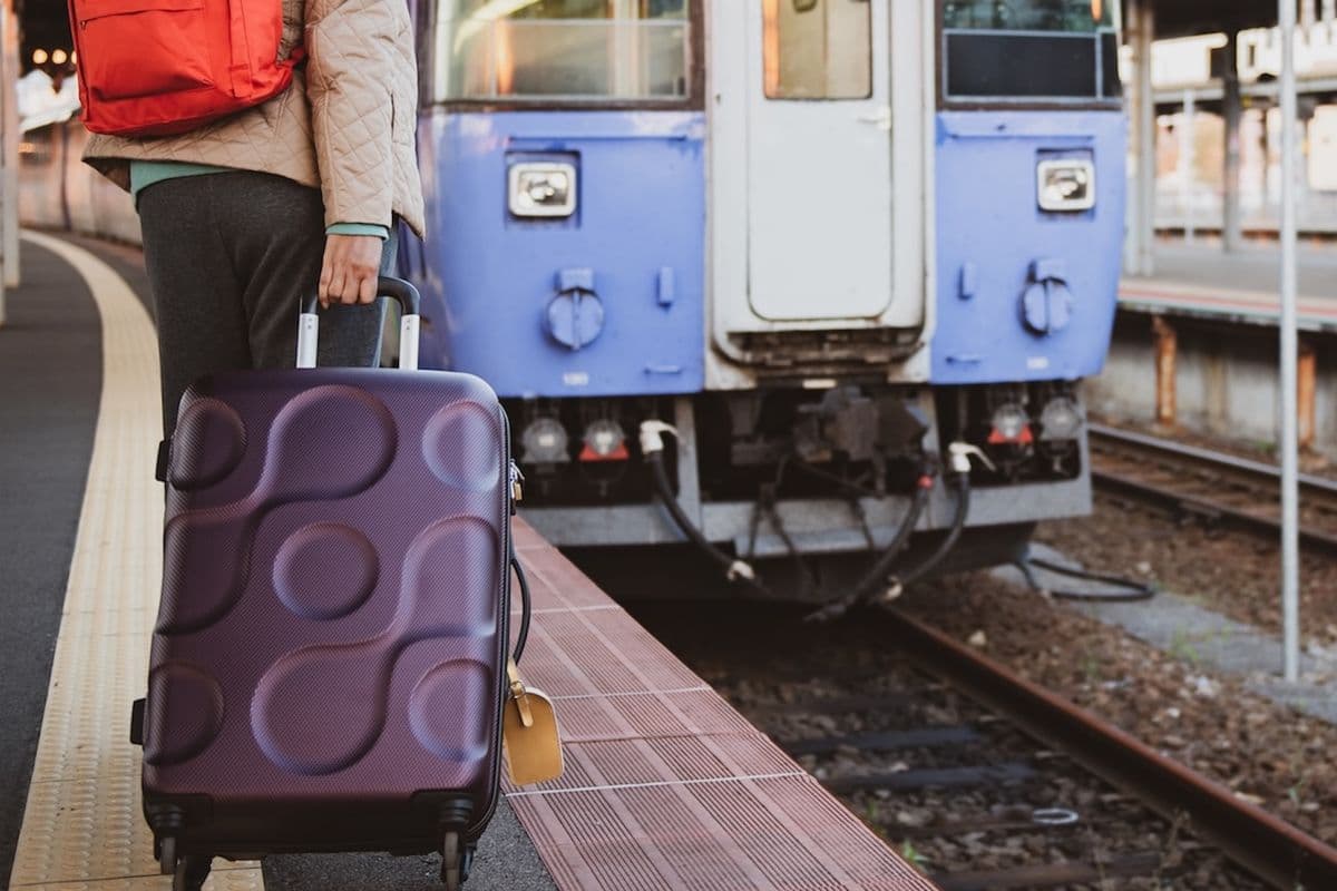 Person with a red backpack and purple suitcase waits at a train station platform beside a blue train.