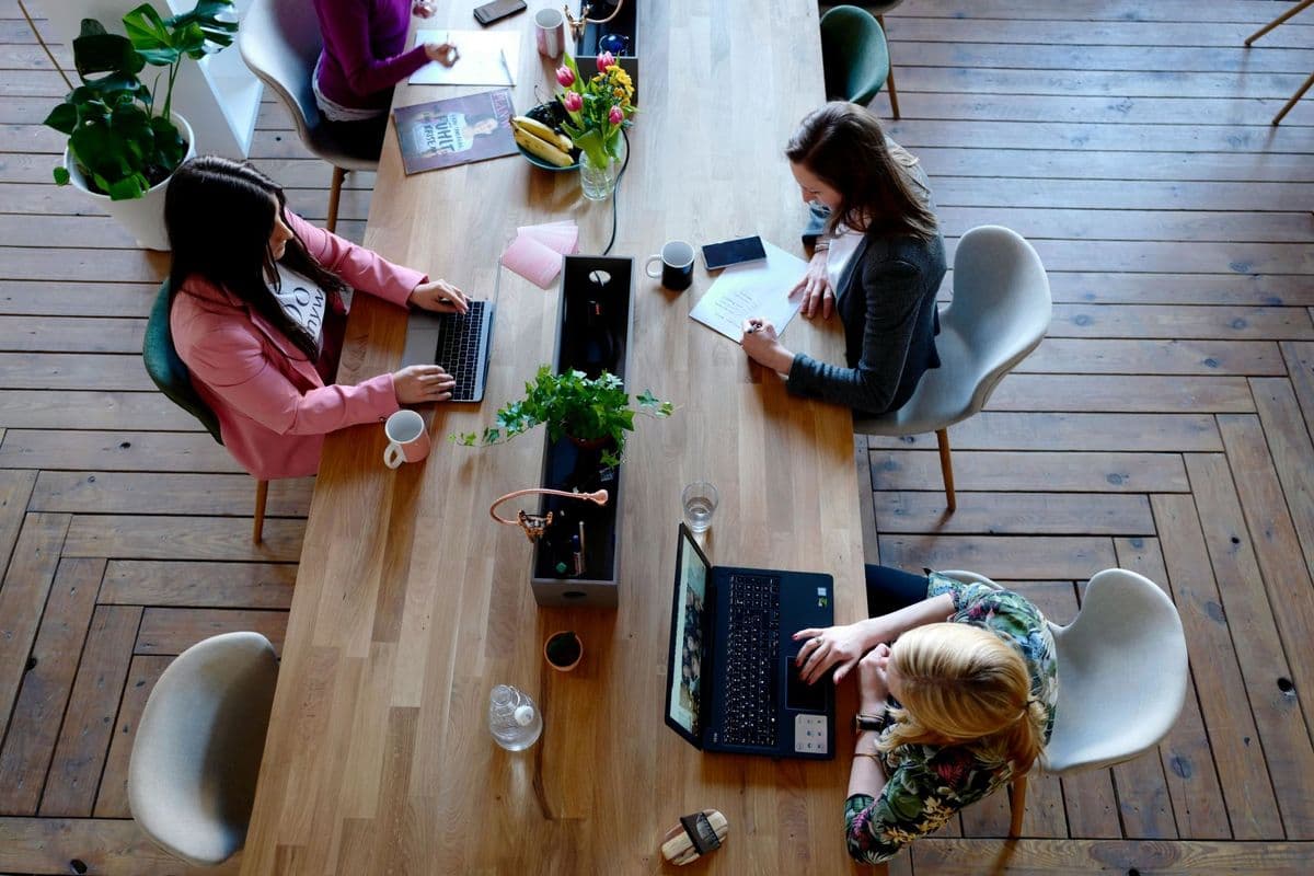 Overhead view of three women working at a wooden table with laptops, notebooks, and coffee, decorated with plants and flowers