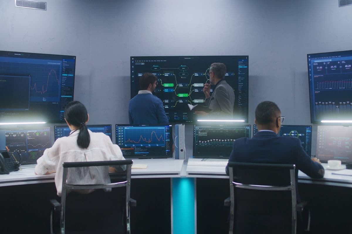 People working in a control room with multiple computer screens displaying data charts and graphs. Two individuals stand discussing by a large screen.